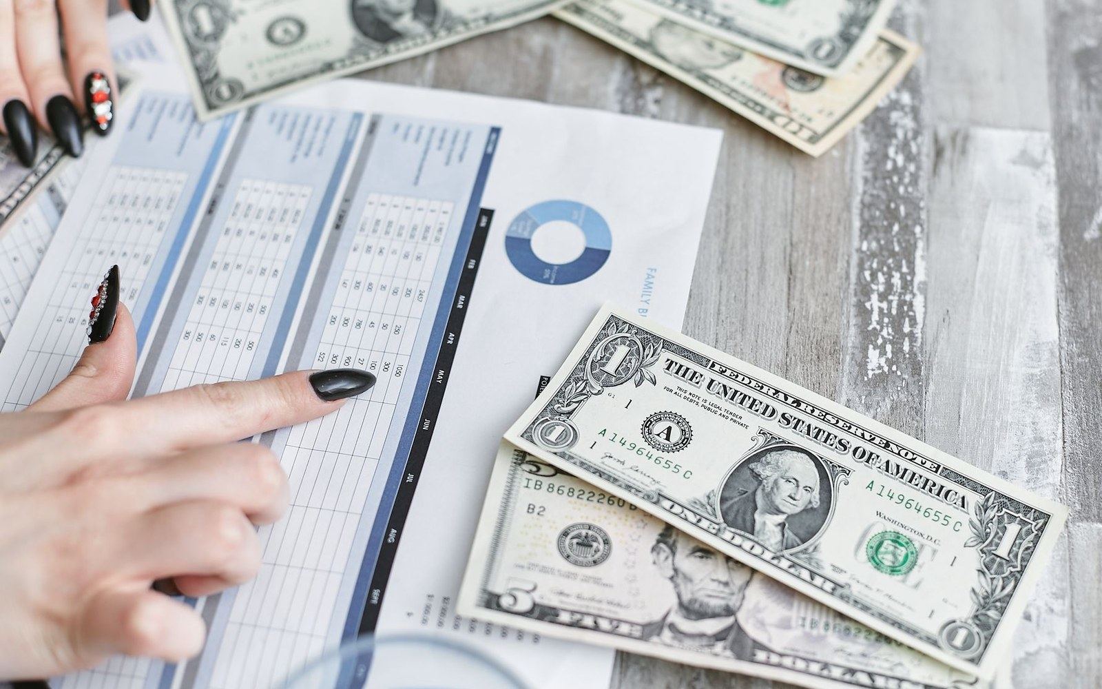 A hand with black nail polish points at a budget spreadsheet beside scattered dollar bills